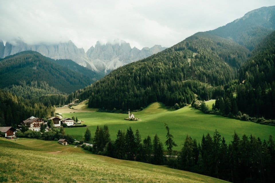 Eine kleine weiße Kirche und ein Dorf liegen in einem lebhaft grünen Tal, umgeben von Pinienwäldern und zerklüfteten Berggipfeln.