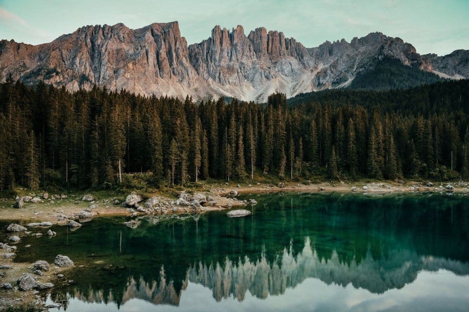 Eine schroffe Bergkette überragt einen dichten Kiefernwald, beides spiegelt sich im stillen, smaragdgrünen Wasser eines Sees.