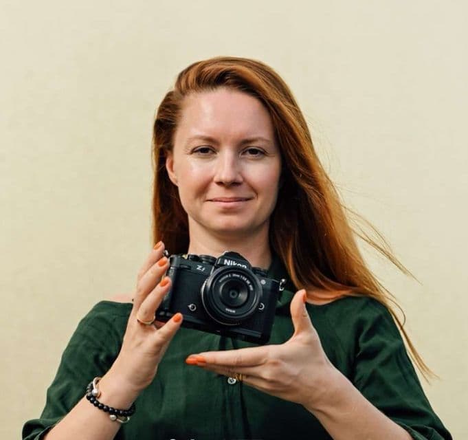 Eine Frau mit langen roten Haaren und einem grünen Oberteil hält eine schwarze Nikon Z spiegellose Kamera mit beiden Händen hoch.