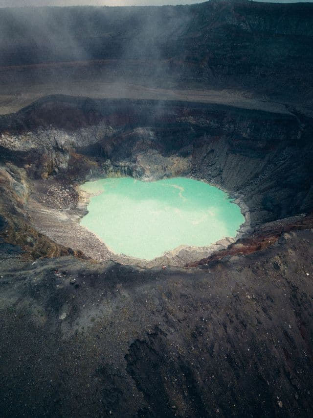 Vista aerea di un lago turchese nel fondo di un grande cratere vulcanico scuro, con vapore che sale dalle pendici.
