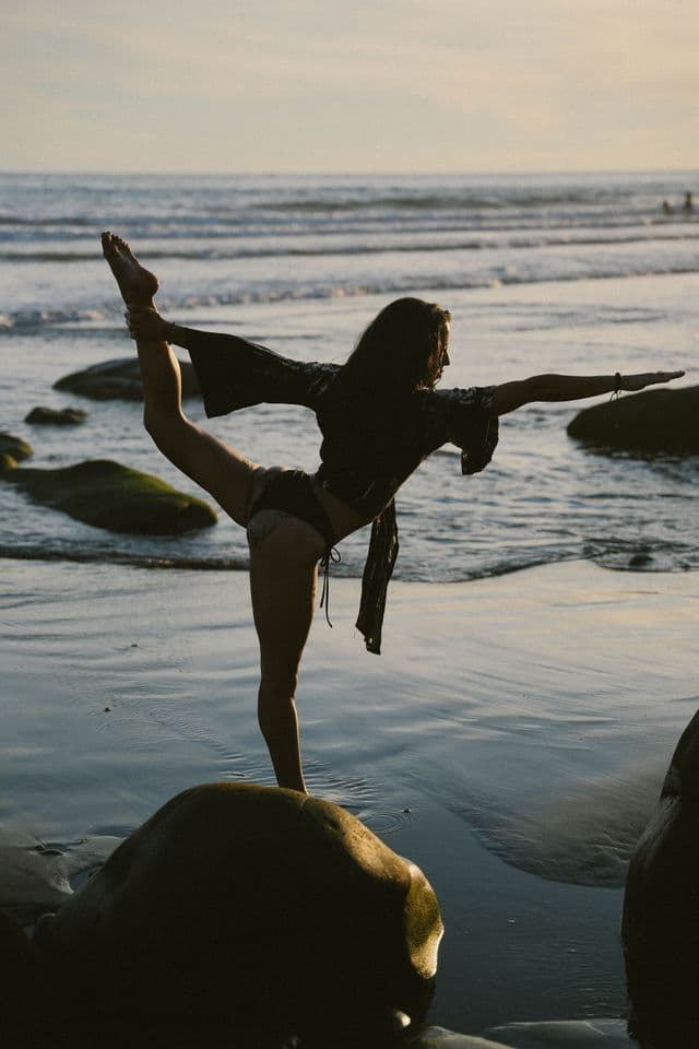 Una donna in silhouette esegue una posa yoga da ballerina nelle acque basse di una spiaggia al tramonto.