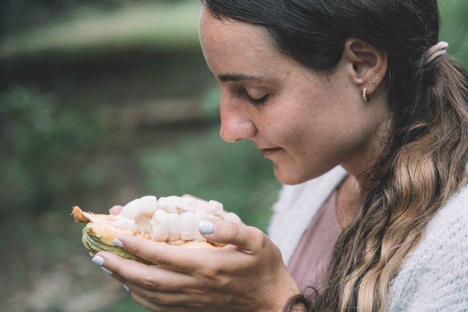 Una donna di profilo tiene un baccello di cacao aperto con polpa bianca, annusandone il contenuto ad occhi chiusi.