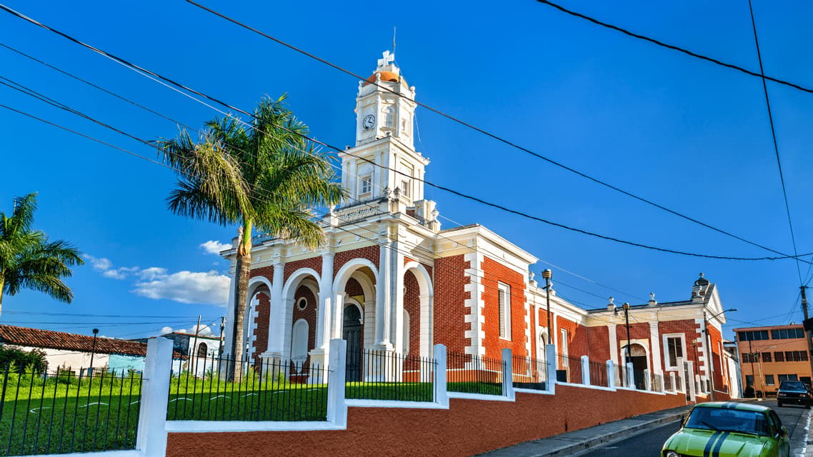 Una chiesa di mattoni rossi e bianca, con campanile, si erge tra le palme sotto un cielo azzurro e limpido, con cavi elettrici aerei.