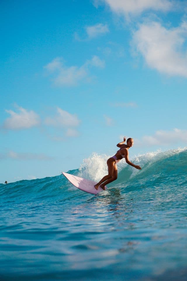 Eine Frau im Bikini surft auf einem pinken Surfbrett und balanciert auf einer sich kräuselnden blauen Welle unter klarem Himmel.