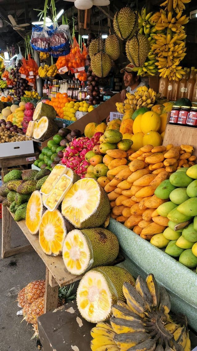 Auf einem Marktstand im Freien türmen sich farbenfrohe tropische Früchte, darunter geschnittene Jackfrucht, Durian, Mangos und Drachenfrucht.