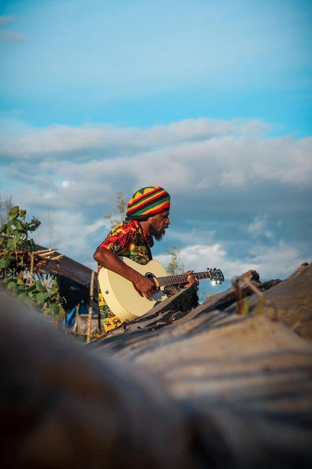 Ein Mann mit bunter Rasta-Mütze und gemustertem Hemd spielt draußen unter bewölktem Himmel eine weiße Akustikgitarre.