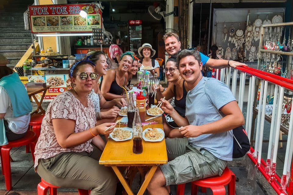 Eine lächelnde WeRoad-Gruppe isst Nudeln mit Stäbchen an einem Außentisch vor einem Streetfood-Stand.