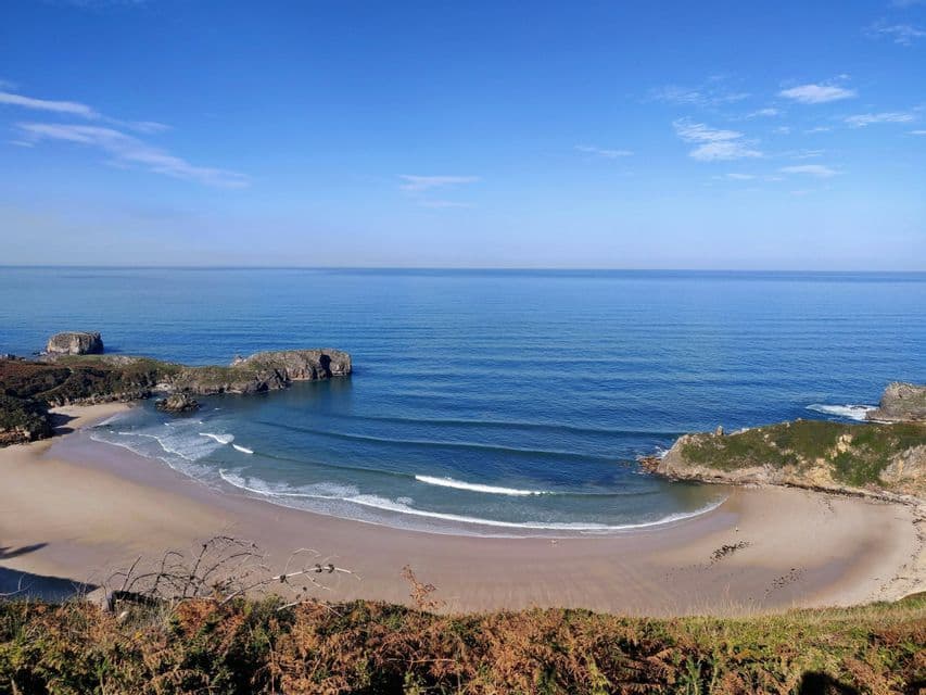 Una vista dall'alto di una spiaggia di sabbia a mezzaluna, con onde dolci che lambiscono la riva di un oceano blu e calmo, sotto un cielo limpido.