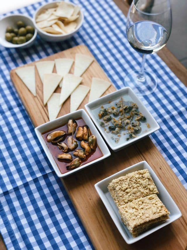 Una tavola imbandita con tapas, formaggio, cozze, olive e un bicchiere di vino bianco su una tovaglia a quadri.
