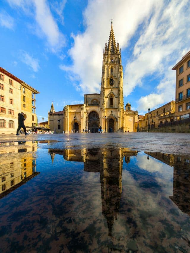 Una cattedrale gotica riflessa in una grande pozzanghera in una piazza cittadina, sotto un cielo blu.