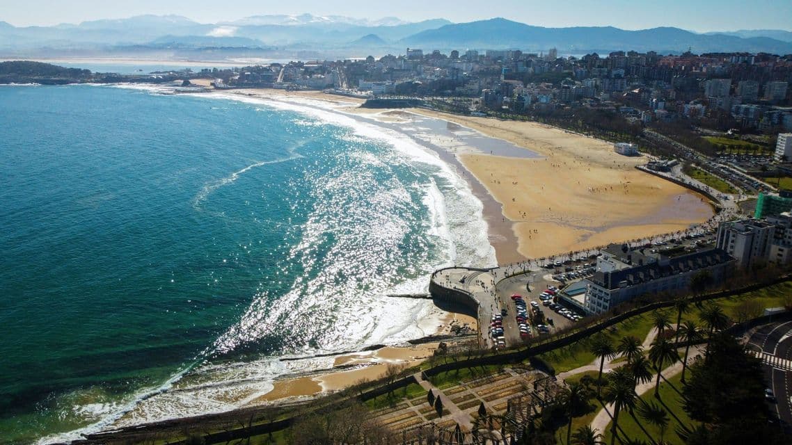 Vista aerea di una città costiera soleggiata con un'ampia spiaggia di sabbia, acqua blu scintillante e montagne in lontananza.