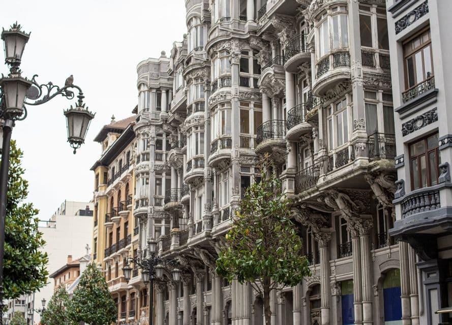 Edifici grigi riccamente decorati con bovindi e balconi dettagliati fiancheggiano una strada cittadina, accanto ad alberi e un lampione d'epoca.