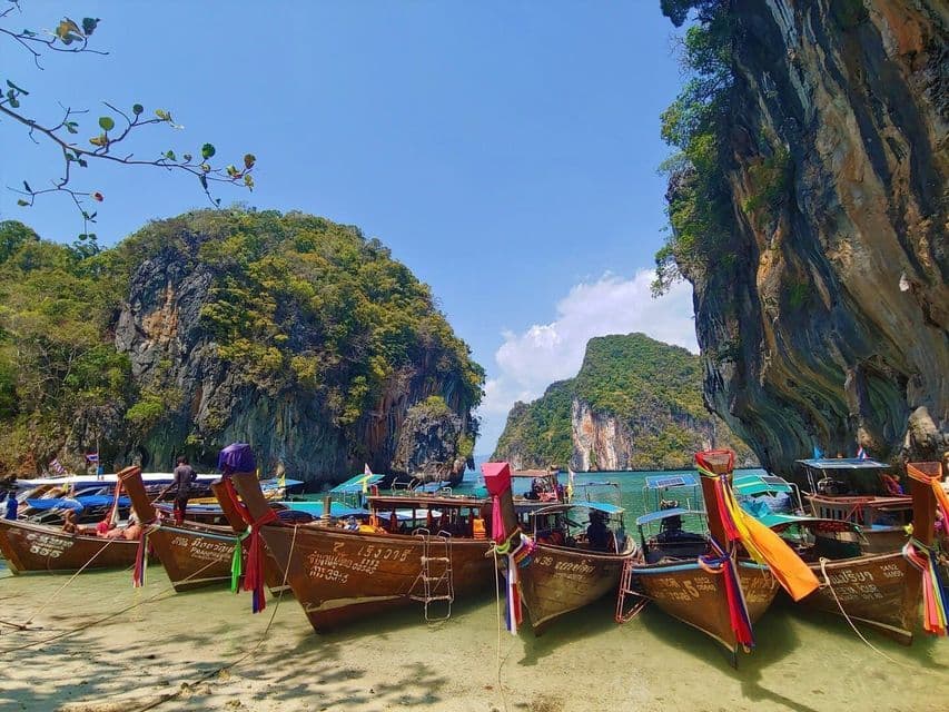 Several traditional long-tail boats with colorful ribbons are docked on a sandy beach in a bay surrounded by steep, green cliffs.