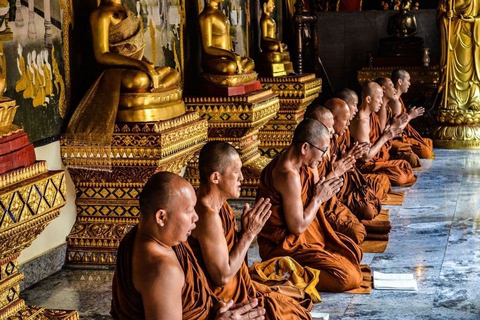 Una fila di monaci buddisti in tuniche arancioni inginocchiati a pregare all'interno di un tempio, con statue dorate di Buddha sullo sfondo.