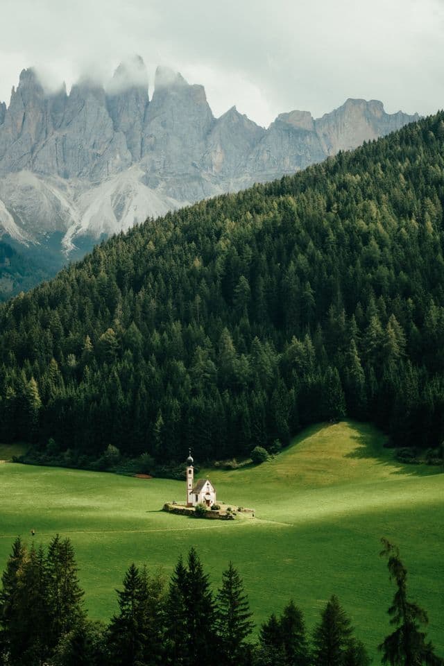 Eine kleine weiße Kirche steht allein auf einer hellgrünen Wiese, mit einem dichten Wald und felsigen Bergen im Hintergrund.