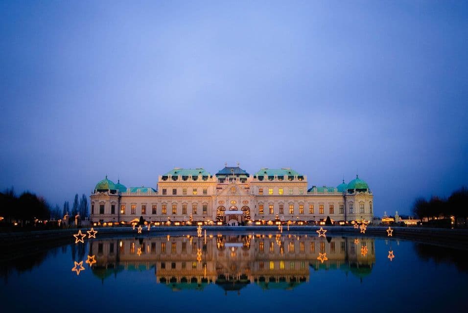 Un palazzo illuminato con cupole verdi che si riflettono perfettamente in un ampio specchio d'acqua al crepuscolo, con decorazioni a stella luminose in primo piano.