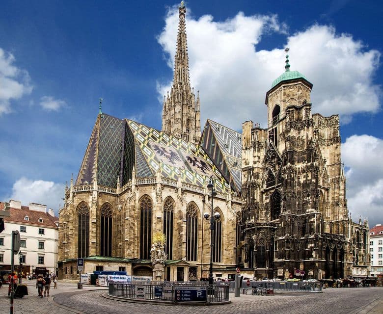 Una grande cattedrale gotica con un tetto in tegole colorate e decorate e un'alta guglia si erge in una piazza acciottolata sotto un cielo azzurro nuvoloso.