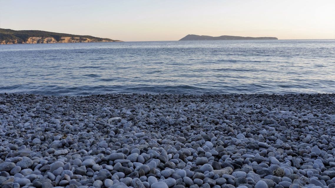 Ein Kiesstrand mit glatten grauen Steinen trifft auf das ruhige Meerwasser, wobei ferne Klippen am Horizont sichtbar sind.