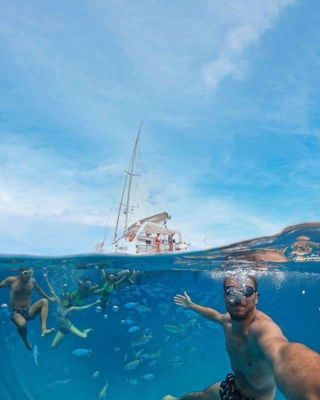 Eine WeRoad Gruppenreise schnorchelt unter Wasser zwischen Fischen, mit einem Mann, der ein Selfie macht, und einem Katamaran an der Oberfläche.