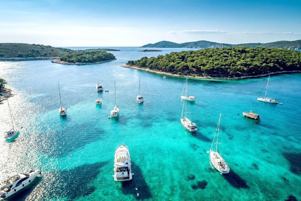 Eine Luftaufnahme von Segelbooten und Yachten, die in einer ruhigen türkisfarbenen Bucht vor Anker liegen, umgeben von grünen, bewaldeten Inseln.