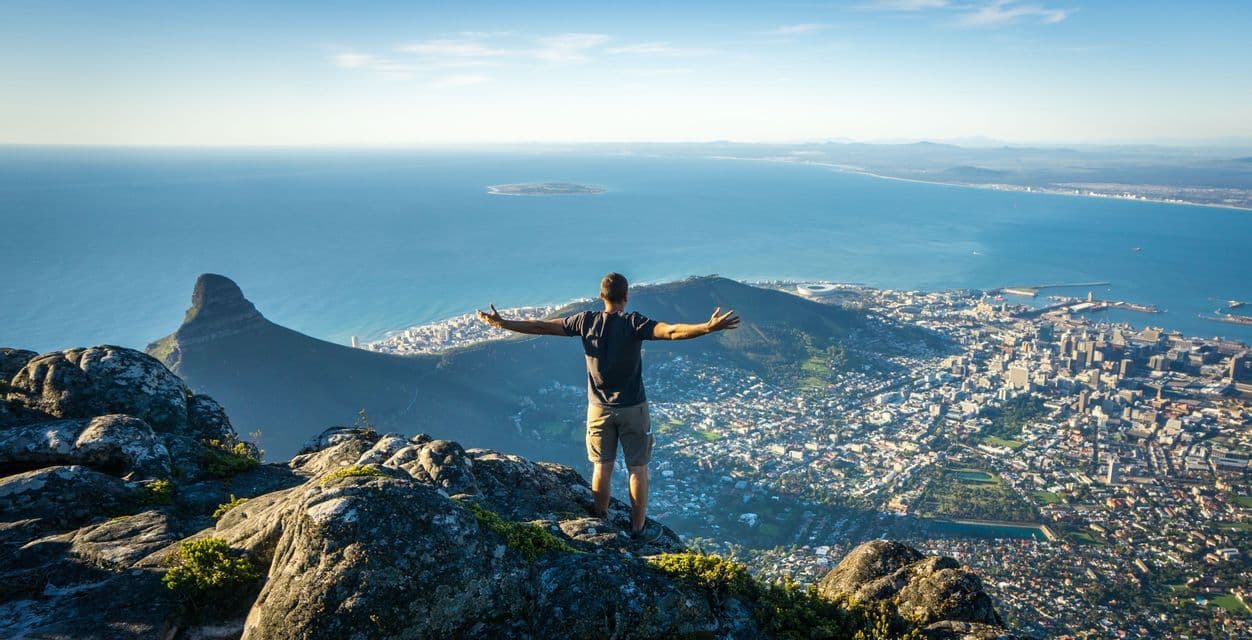 A person stands on a rocky mountain summit with arms outstretched, overlooking a coastal city and the ocean.