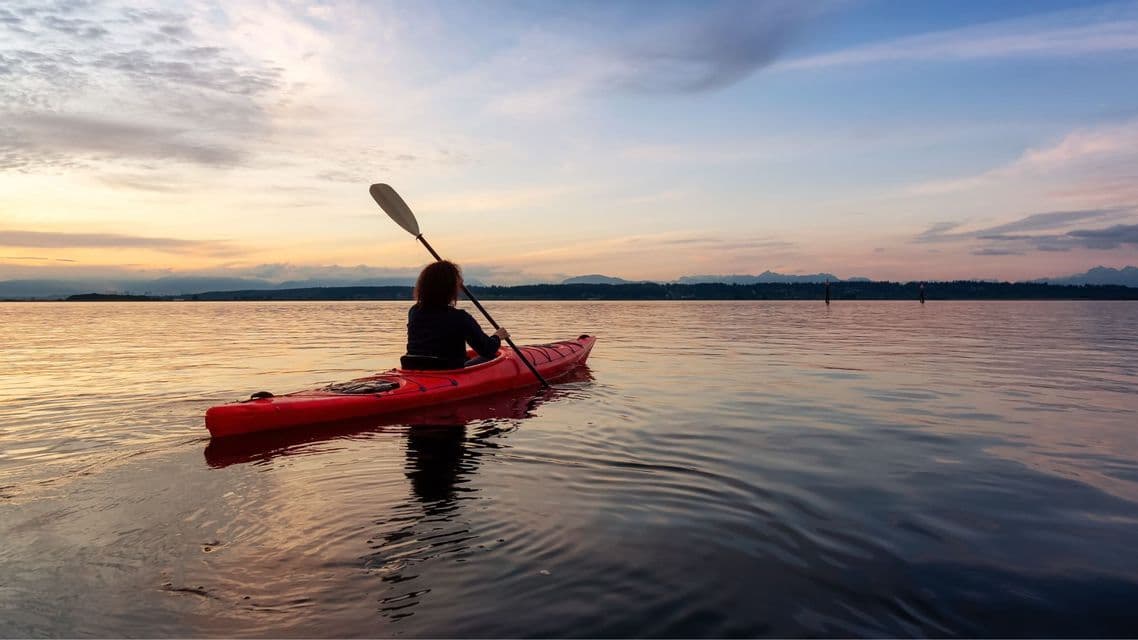 Una persona in un kayak rosso rema su acque tranquille, osservando il tramonto su una costa lontana.