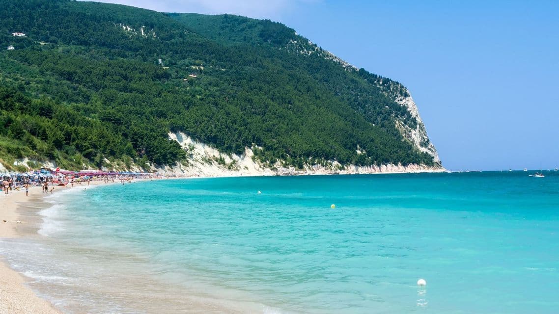 Una spiaggia sabbiosa affollata incontra acque turchesi ai piedi di una grande scogliera alberata sotto un cielo azzurro e limpido.