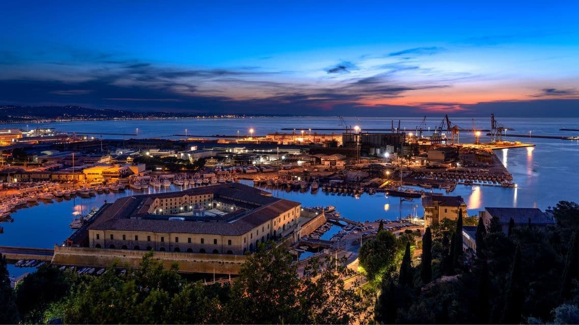 Una città portuale illuminata e il suo porto al tramonto, con un grande edificio storico e barche ormeggiate.