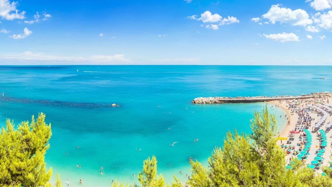 Una veduta aerea di una spiaggia affollata con molti ombrelloni, gente che nuota nel mare turchese e un molo in pietra sotto un cielo azzurro.