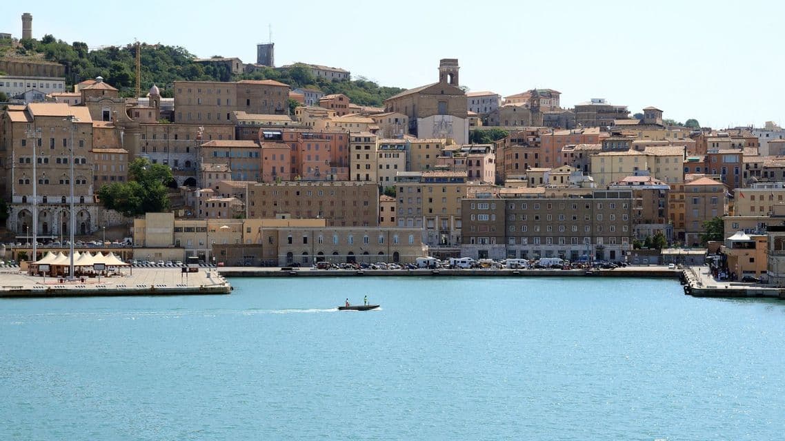 Una piccola imbarcazione naviga in un porto, di fronte a una città densa con edifici color sabbia e rosa costruiti su un pendio verde.