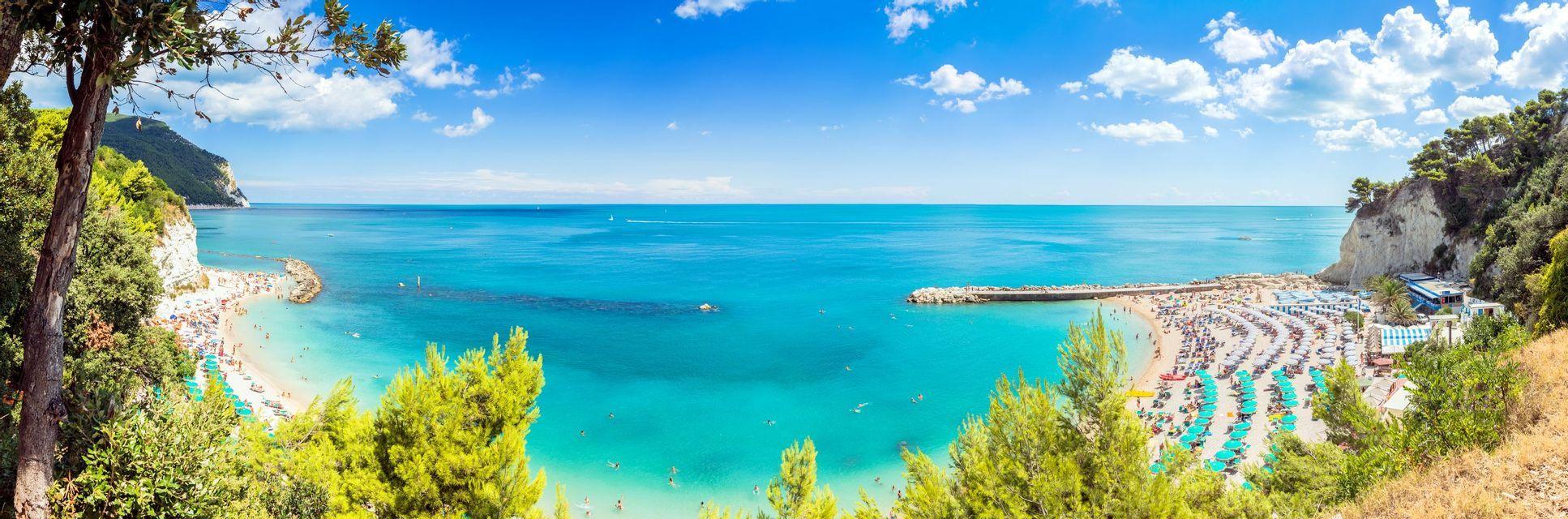 Vista panoramica di una spiaggia affollata con acqua turchese, incastonata in una cala tra scogliere verdi sotto un cielo azzurro.