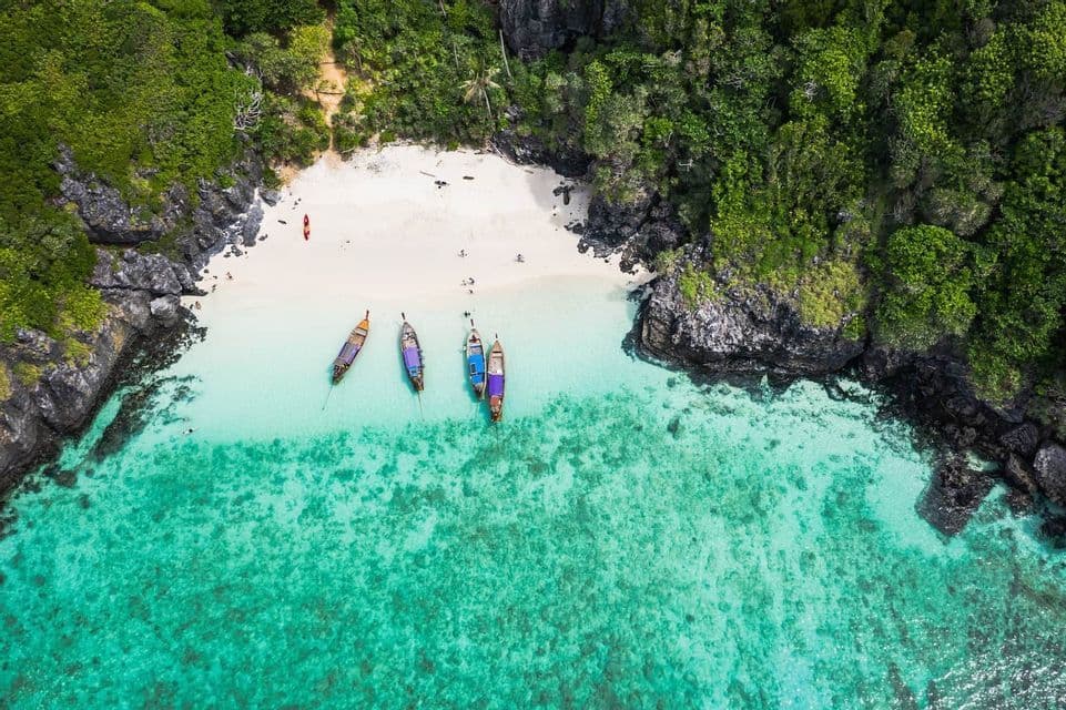 Vista aerea di quattro barche a coda lunga ancorate in acque limpide color turchese vicino a una spiaggia isolata di sabbia bianca e scogliere ricoperte di giungla.