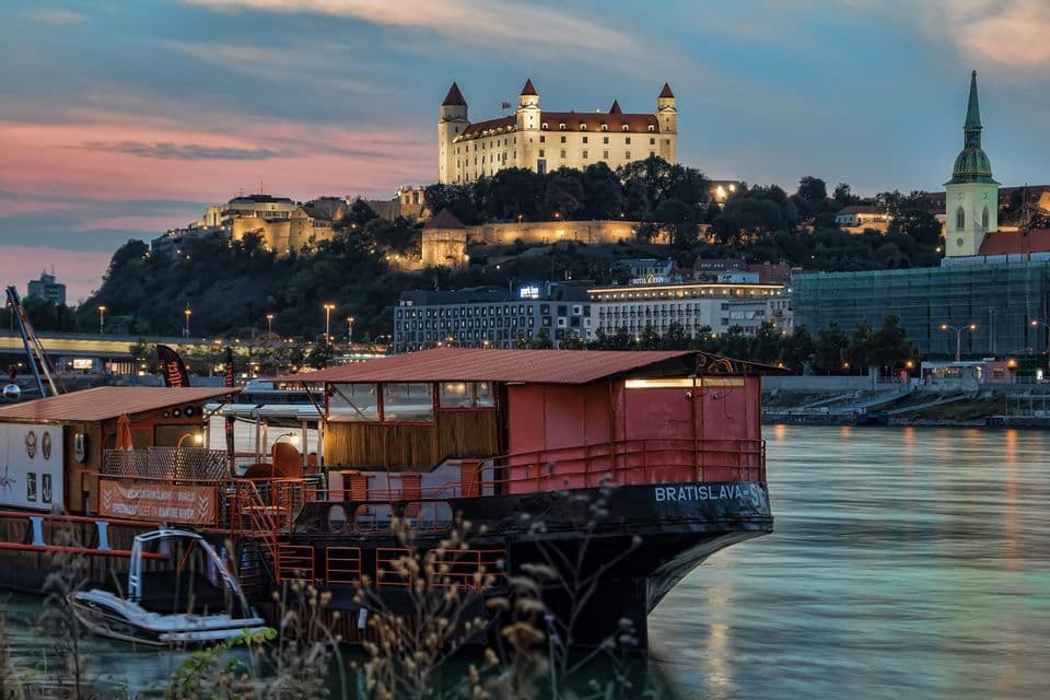 Una barca rossa attraccata su un fiume, con un castello illuminato su una collina sullo sfondo al crepuscolo.