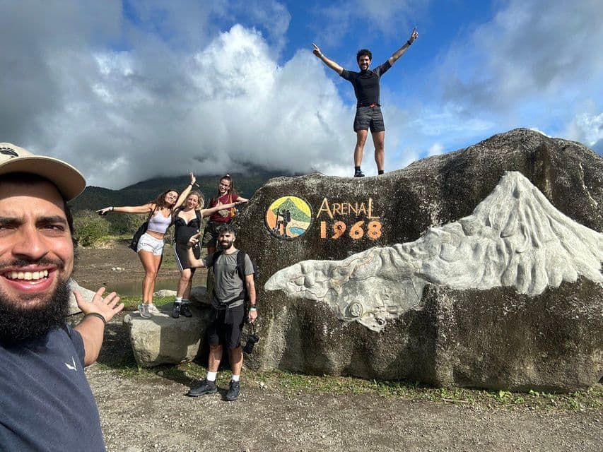 Un groupe WeRoad prend un selfie devant un grand rocher sculpté avec "Arenal 1968" et un relief de volcan.