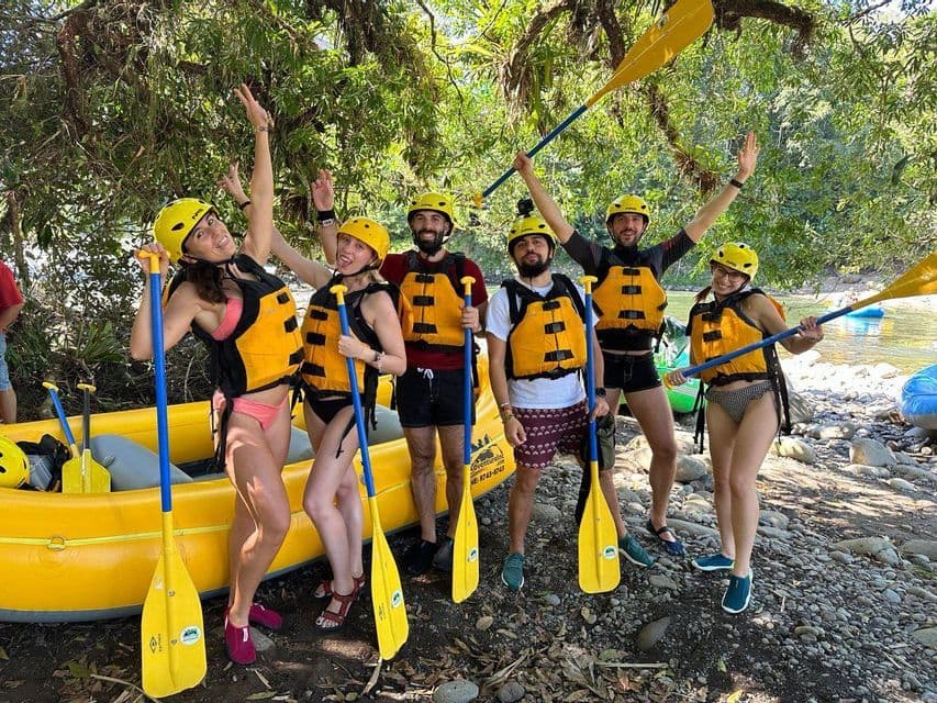 A WeRoad group trip in helmets and life vests posing with paddles next to a raft on a riverbank.