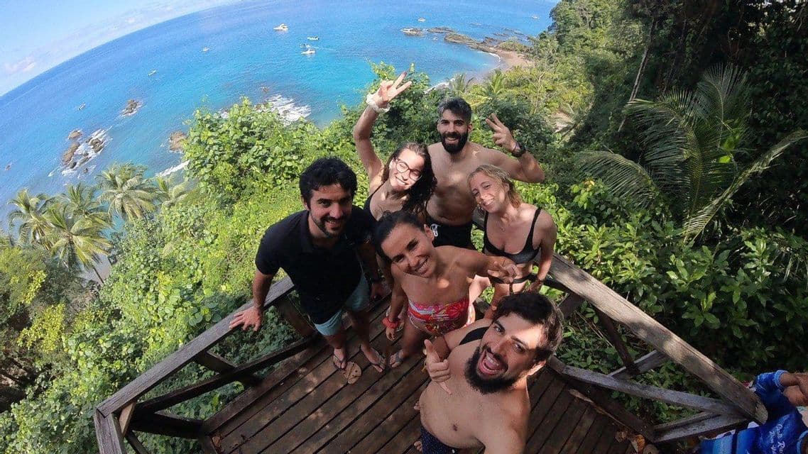 Un groupe WeRoad prend un selfie souriant sur un point de vue en bois surplombant une côte tropicale aux eaux bleues.