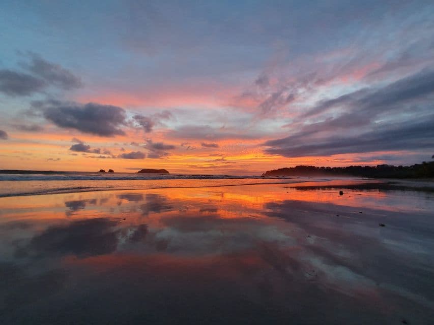 A vibrant sunset with orange, pink, and blue clouds reflecting on the mirror-like wet sand of a beach.