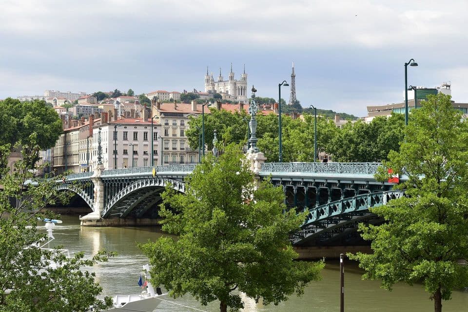 Un elaborato ponte verde attraversa un fiume, conducendo a un panorama cittadino con una cattedrale e una torre in cima a una collina, sotto un cielo nuvoloso.