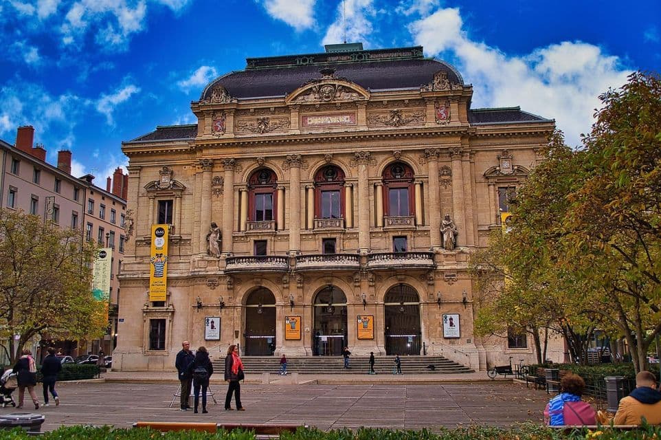 Un sontuoso edificio teatrale in stile classico si erge in una piazza cittadina, con persone che passeggiano sotto un cielo azzurro.