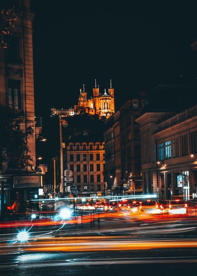 Una fotografia a lunga esposizione di una strada cittadina di notte, con scie luminose di auto e una basilica illuminata su una collina.