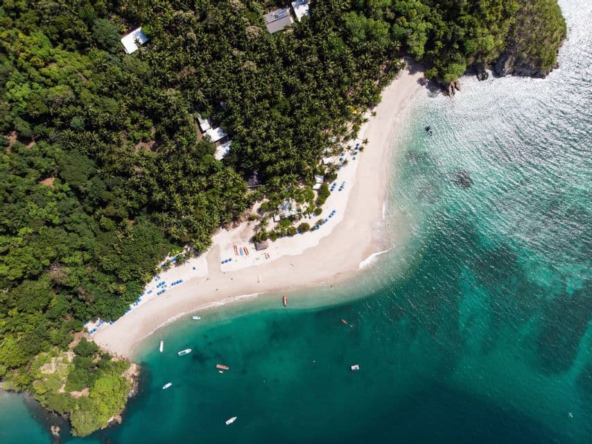 Una vista aérea cenital de una playa de arena blanca que bordea una exuberante selva de palmeras, con barcos flotando en agua turquesa cristalina.