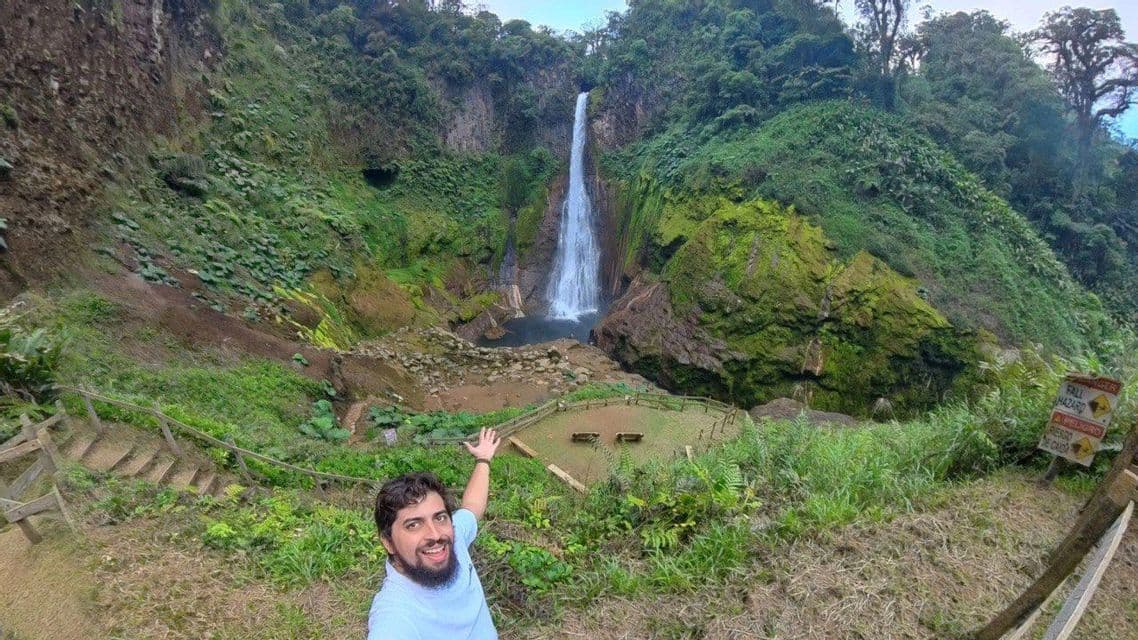 Un homme souriant et barbu prend un selfie en désignant une haute cascade dans une vallée luxuriante et verte.