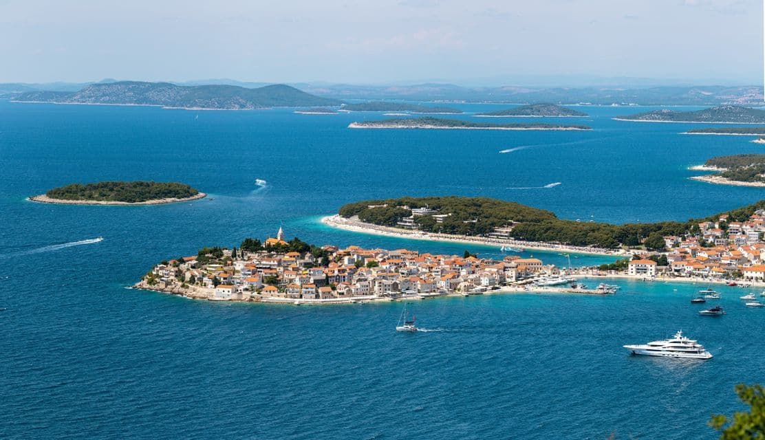 Vista aerea di una cittadina costiera su una piccola penisola, circondata da un mare blu intenso, barche e un arcipelago di isole verdi.