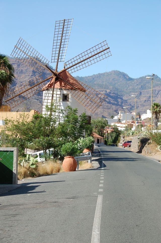 Un tradizionale mulino a vento bianco con tetto in terracotta sorge lungo una strada, con montagne e un villaggio sullo sfondo.