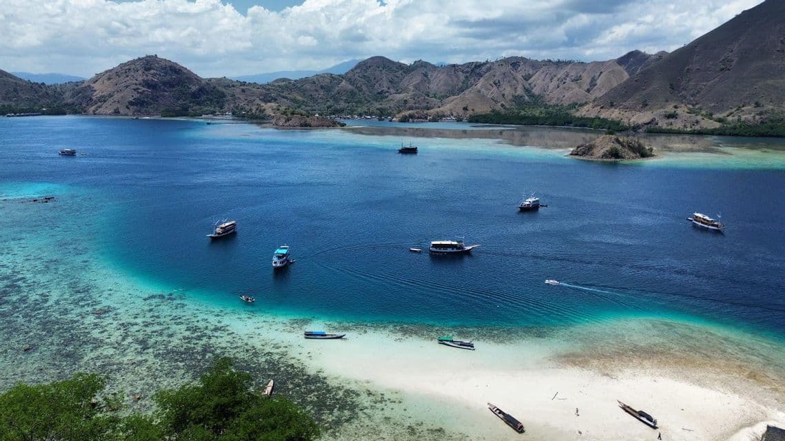 Vue aérienne d'une baie tropicale avec des bateaux sur une eau turquoise, une plage de sable blanc et des îles vallonnées en arrière-plan.