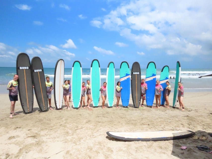 Un groupe WeRoad posant avec leurs planches de surf sur une plage de sable, l'océan en arrière-plan.
