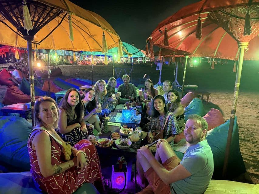 Un groupe WeRoad est assis sur des poufs autour d'une table sur une plage de sable, la nuit, sous des parasols éclairés et colorés.