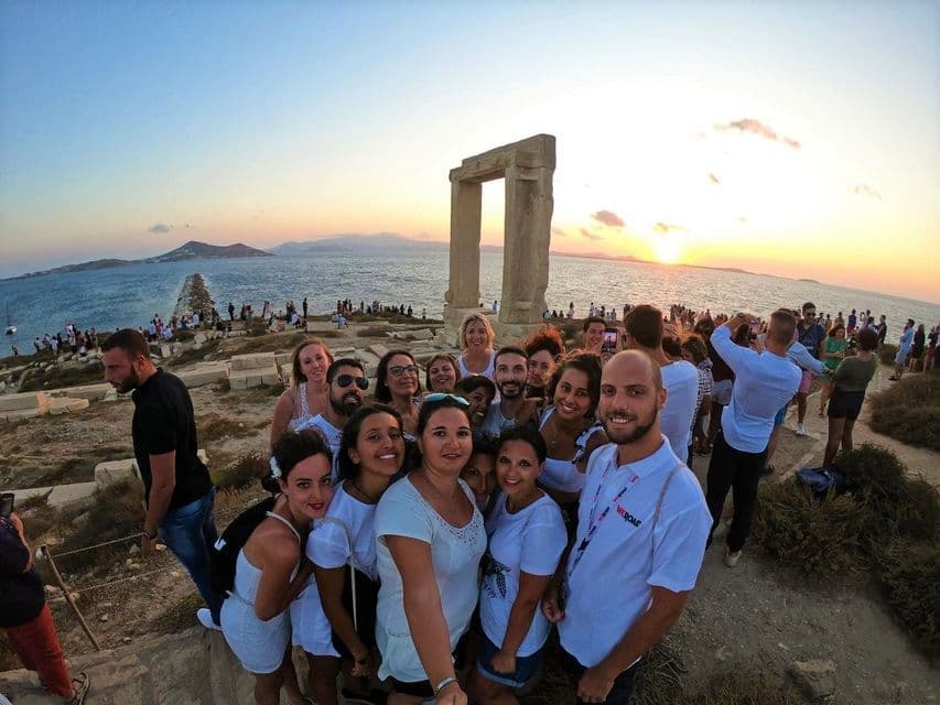 Eine WeRoad Gruppe macht ein Selfie vor einem großen Steinbogen am Meer bei einem farbenprächtigen Sonnenuntergang.