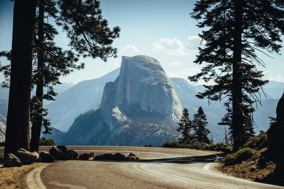 Une vue d'un grand pic de granite depuis une route sinueuse, encadrée de hauts pins sous un ciel bleu clair.
