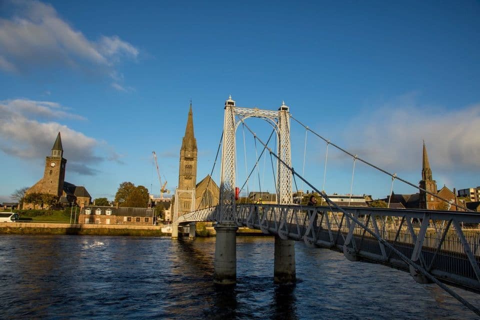 Eine weiße Hängebrücke überspannt einen Fluss mit historischen Kirchen und Gebäuden am Ufer unter blauem Himmel.